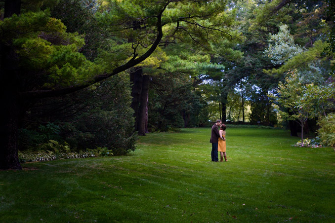 Engaged couple in open, manecured lawn with trees towering around the edges